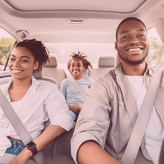 Casal e criança negros sorrindo dentro de um carro. O casal nos bancos da frente e a criança atrás.