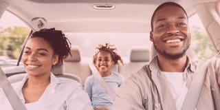 Casal e criança negros sorrindo dentro de um carro. O casal nos bancos da frente e a criança atrás.