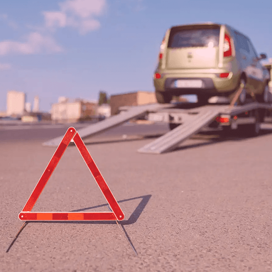 Triângulo de sinalização colocado no chão de uma rua. Ao fundo, um carro em cima de um guincho.