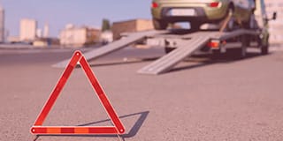 Triângulo de sinalização colocado no chão de uma rua. Ao fundo, um carro em cima de um guincho.