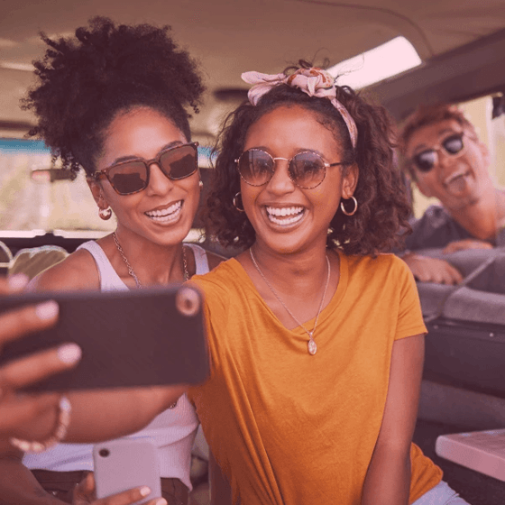 Duas mulheres jovens negras sorrindo dentro de um carro. Na parte de trás, um homem branco está para fora do carro entrando com a cabeça pela janela sorrindo. Os três estão posando para uma selfie no celular.