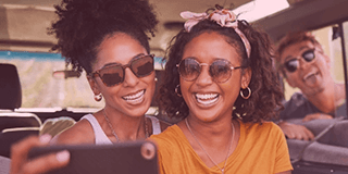 Duas mulheres jovens negras sorrindo dentro de um carro. Na parte de trás, um homem branco está para fora do carro entrando com a cabeça pela janela sorrindo. Os três estão posando para uma selfie no celular.