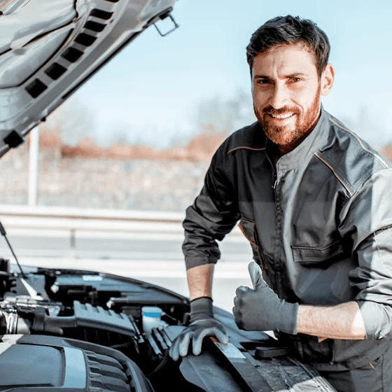 Homem de uniforme de mecânico sorrindo com capô do carro aberto.