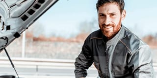 Homem de uniforme de mecânico sorrindo com capô do carro aberto.
