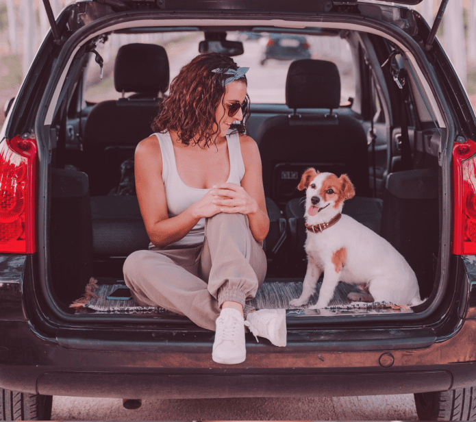 Mulher de óculos escuros sentada com o cachorro de porte pequeno no porta mala do carro